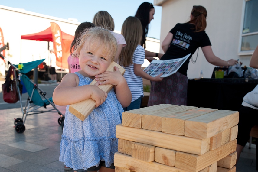Photo of a toddler hugging a wooden block at a community event in the Kāpiti Lights area, with adults and older children in the background