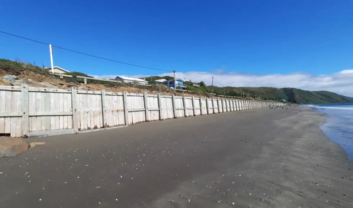 A long stretch of the timber seawall at Paekākāriki, shown from the beach looking north, with weathered wooden panels, dunes and houses above the wall, and a calm shoreline under a bright blue sky.