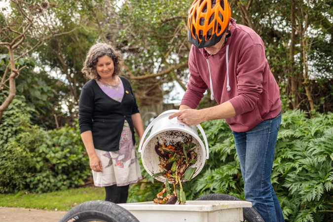 Photo of a community member from Paekākāriki wearing a cycle helmet, picking up compost and putting it into a bike trailer, while a woman watches on.