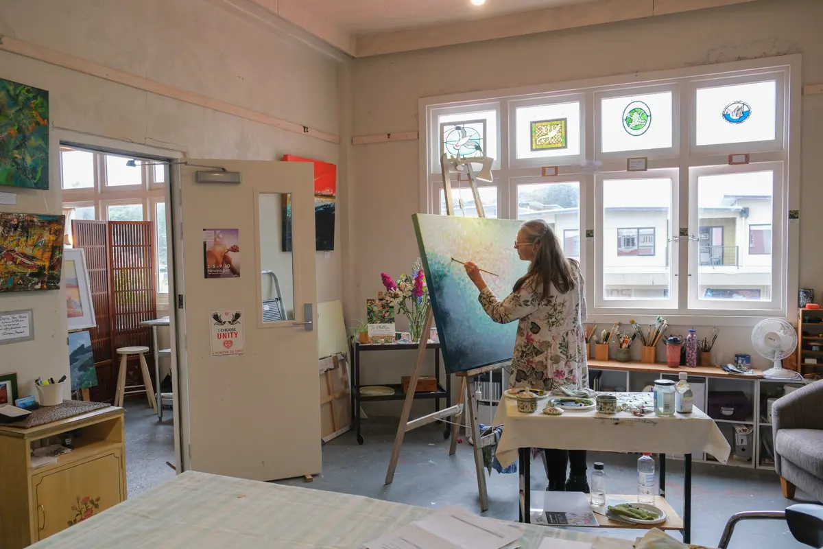 Artist painting a large canvas on an easel inside a light-filled studio. Finished artworks hang on the walls, while brushes, paints and other art materials are arranged on tables beneath stained-glass window panels.