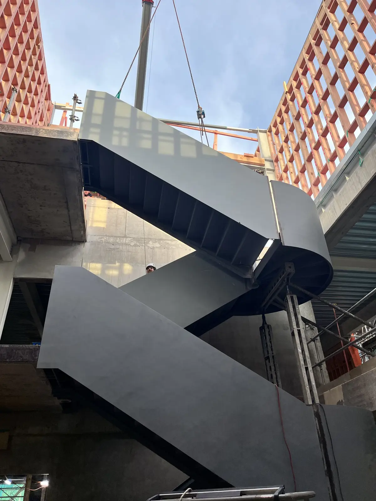 A large dark-grey steel staircase is being lowered into place by a crane inside a multi-storey building under construction. The staircase features two switchback flights with a curved landing and is surrounded by exposed concrete floors, timber wall framing, scaffolding, and construction supports. Open sky is visible above the structure.