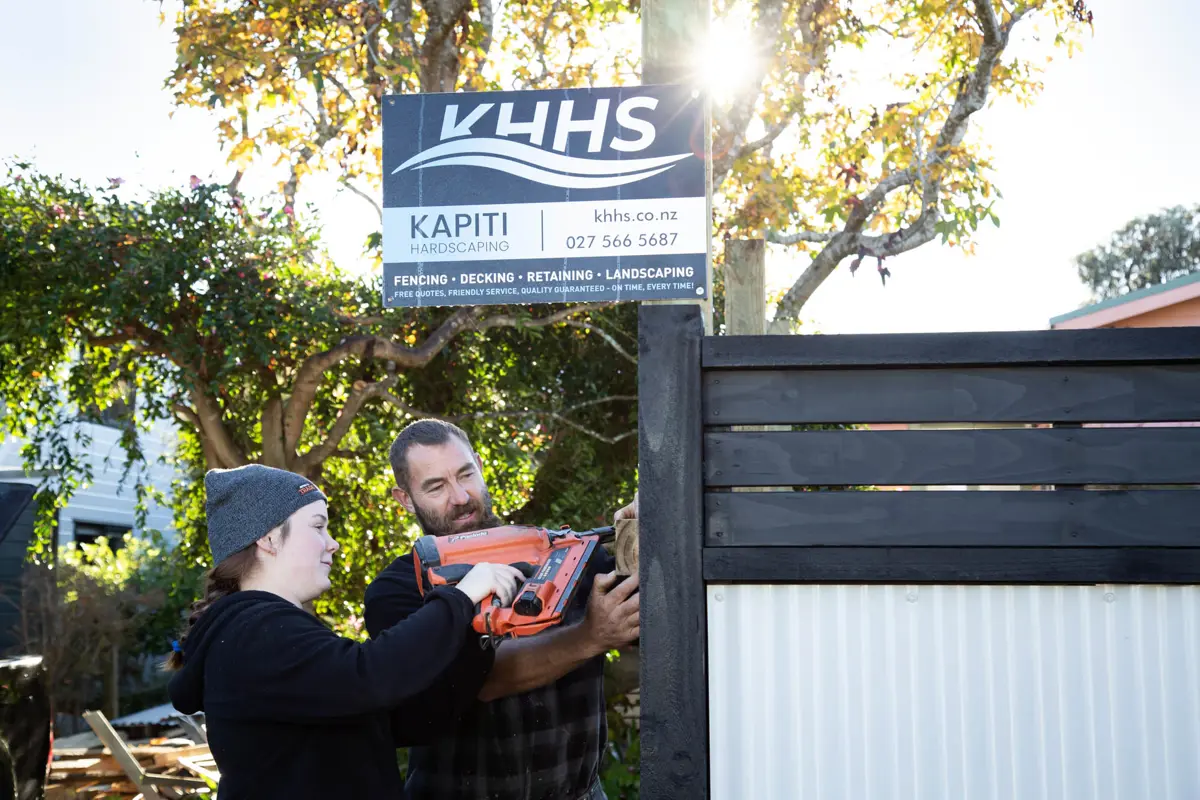 Two people install fencing using a nail gun beside a sign reading “KHHS Kāpiti Hardscaping,” with trees and sunlight in the background.