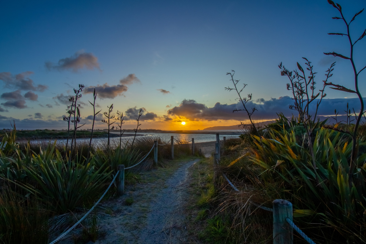 Photo of sunset off the south of Kāpiti Island, looking across the Waikanae River mouth, with a track and flax in the foreground.