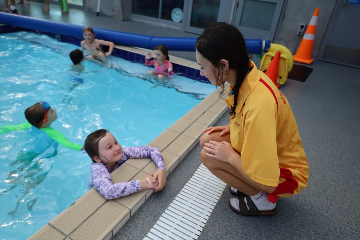 Young lifeguard crouched down pool side talking to a young swimmer in the pool looking up smiling.