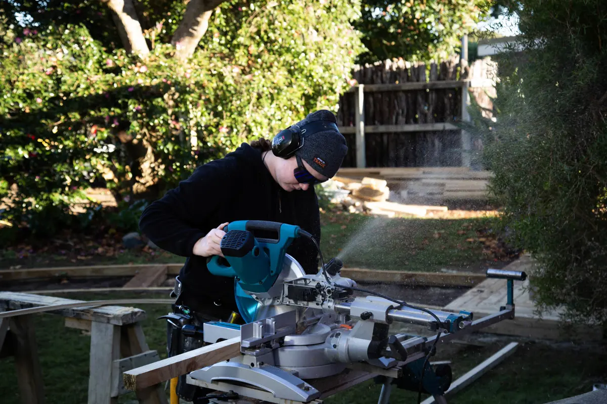 Person operates a drop saw cutting timber outdoors, wearing hearing and eye protection, with sawdust spraying and garden landscaping elements in the background.