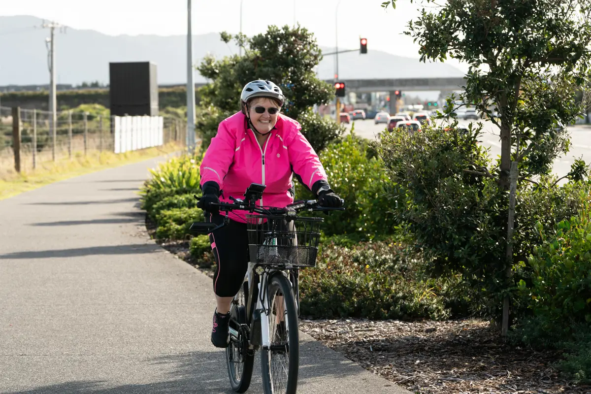 A person wearing a helmet and bright pink jacket rides a bicycle along a shared path bordered by trees and low shrubs, with a road, traffic lights, and cars visible in the background.