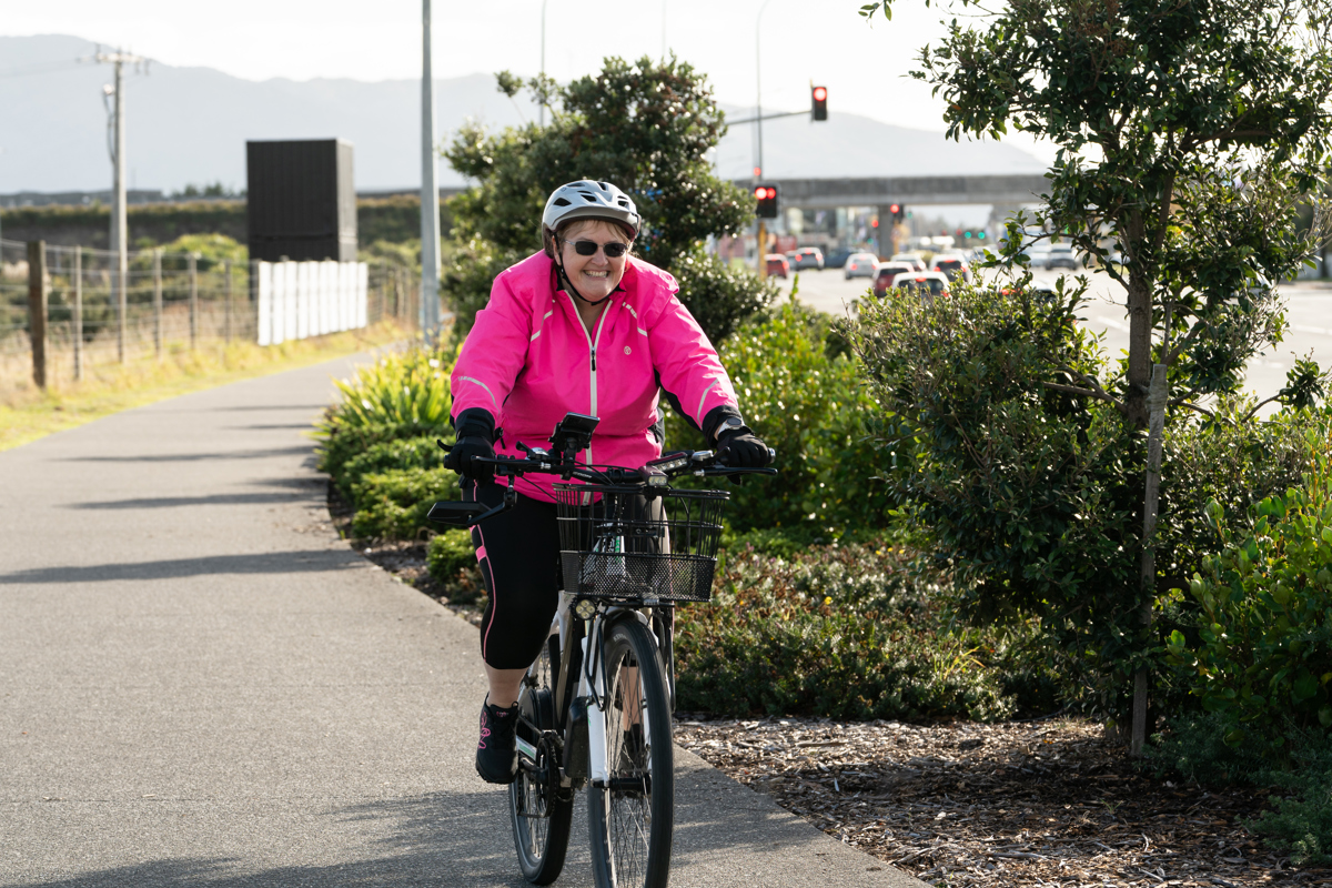 A person wearing a helmet and bright pink jacket rides a bicycle along a shared path bordered by trees and low shrubs, with a road, traffic lights, and cars visible in the background.