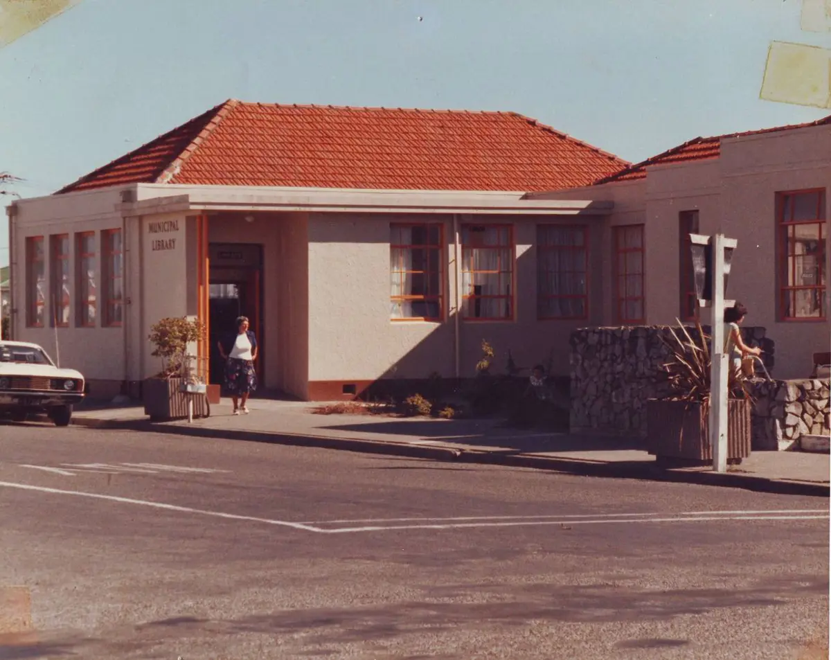 A vintage photo of a municipal library building with a red-tiled roof and light-colored exterior, viewed from across a street. A woman stands near the entrance, and another person is visible near a stone wall on the right. A car is parked on the left side of the building. The scene is sunny with long shadows and a quiet suburban street atmosphere.
