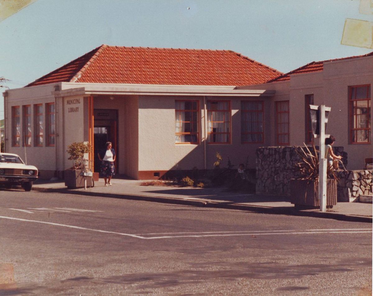 A vintage photo of a municipal library building with a red-tiled roof and light-colored exterior, viewed from across a street. A woman stands near the entrance, and another person is visible near a stone wall on the right. A car is parked on the left side of the building. The scene is sunny with long shadows and a quiet suburban street atmosphere.
