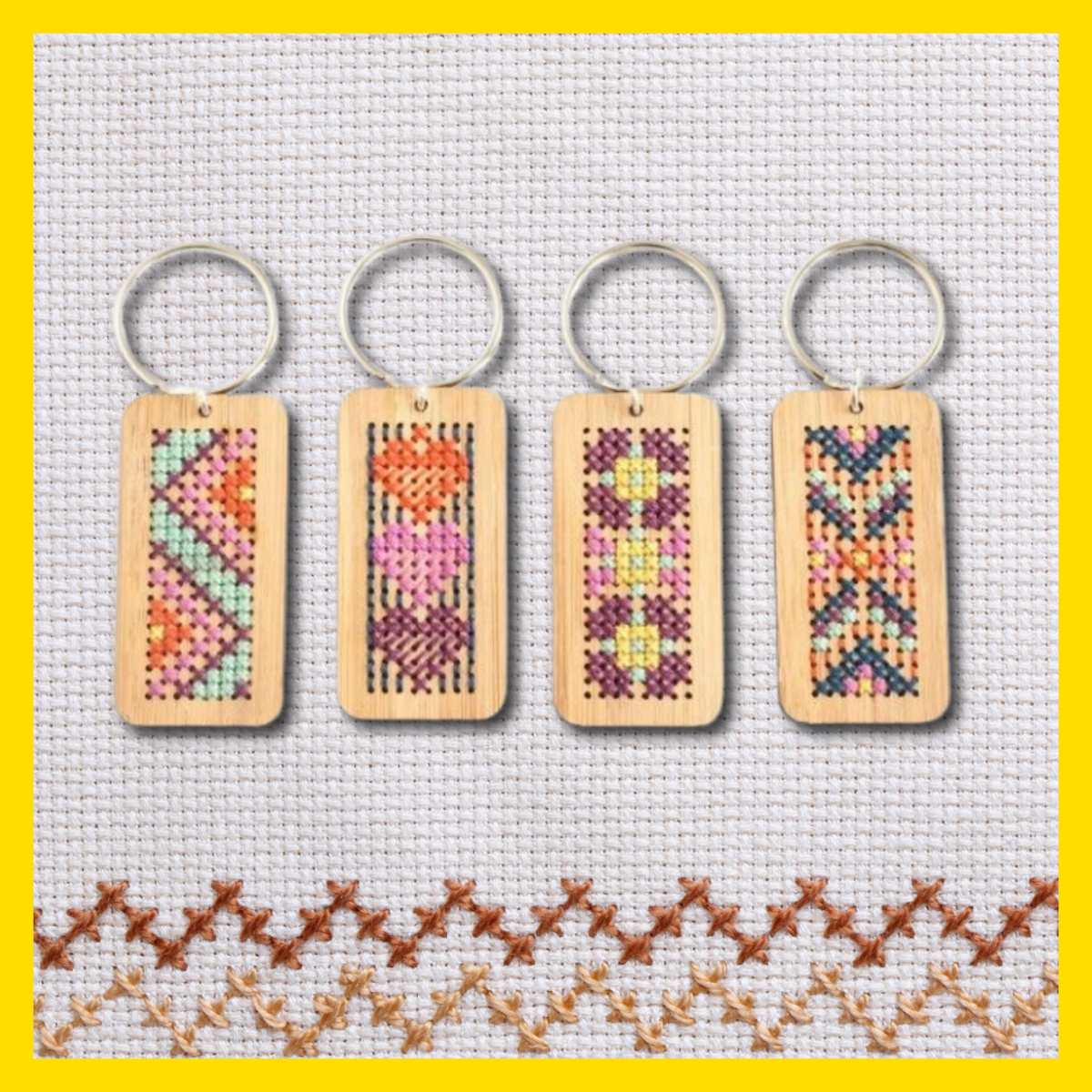 Four wooden rectangular keychains with colourful cross-stitch patterns are displayed on a white cross-stitch fabric background. Each keychain features a different design, including geometric shapes, hearts, flowers, and decorative motifs, attached to silver key rings. A yellow border frames the image.