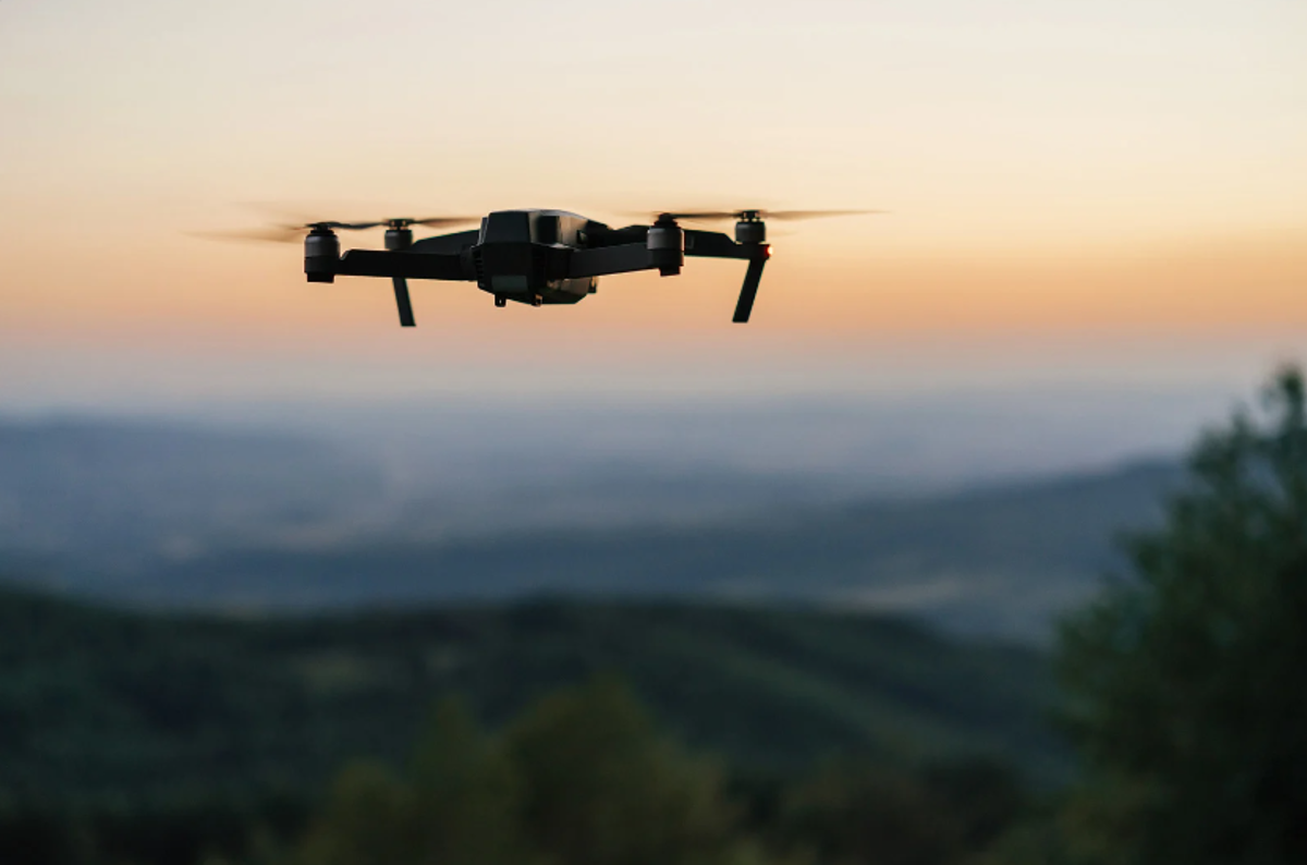Photo of a drone flying against a blurred sunset or sunrise background.