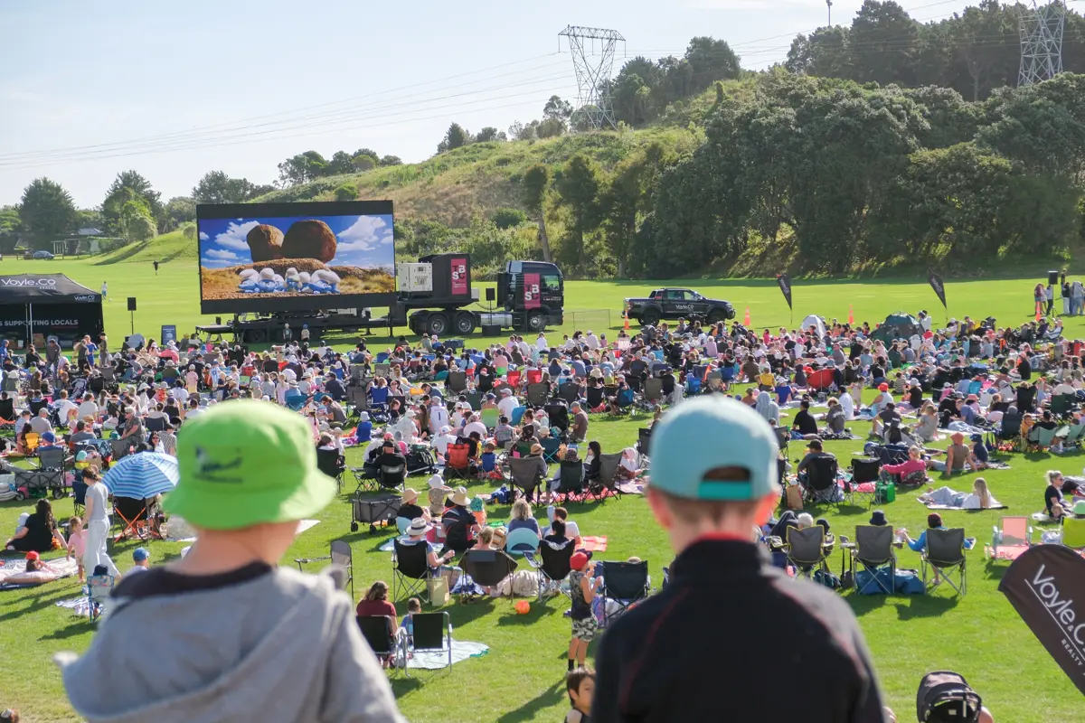 Two boys in the foreground sitting on a hill at movies in the park looking out to a crowd of people in background and a big screen with Smurfs playing