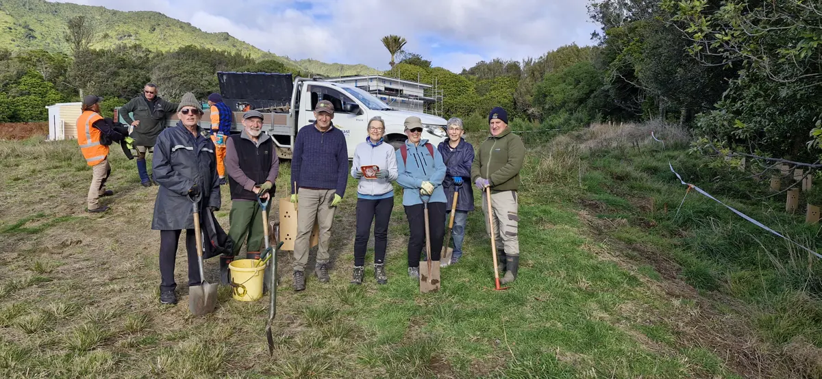 A group of volunteers stand in a grassy outdoor area holding spades and tools, with a work truck and others preparing equipment behind them, and green hills and trees in the background.