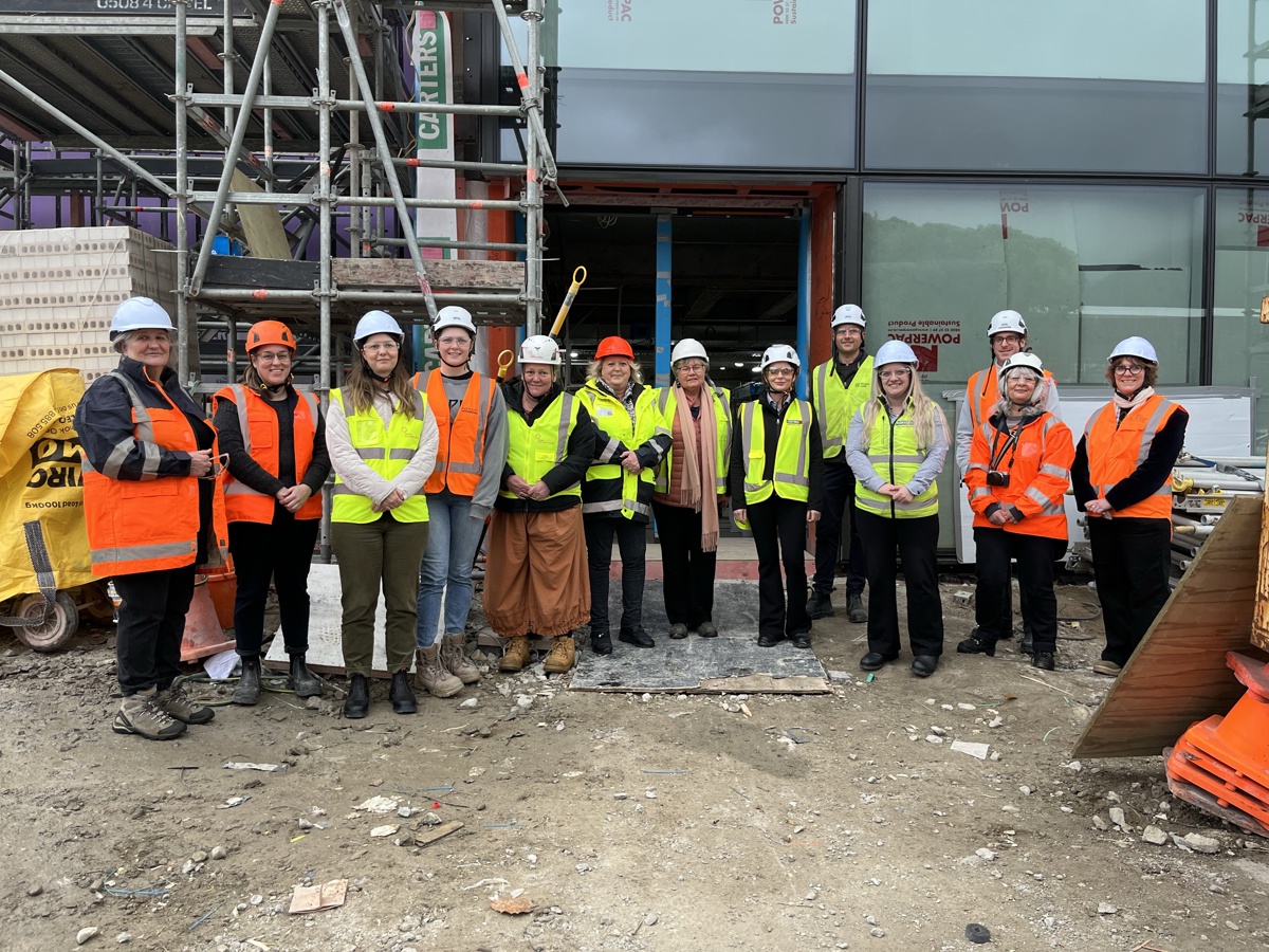 Group of people wearing hard hats and high-visibility clothing stand together for a photo at a construction site. Scaffolding, building materials, and the partially completed exterior of a large building can be seen behind them.
