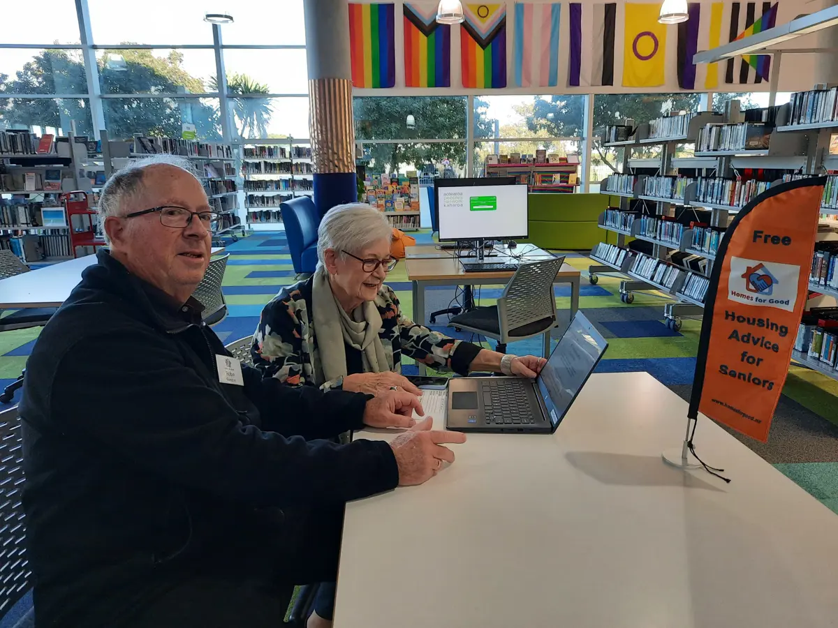 Two older adults sit at a table in Paraparaumu Library using a laptop, with bookshelves, study tables, and rainbow pride flags in the background. An orange table sign reads “Free housing advice for seniors” with the Homes for Good logo.
