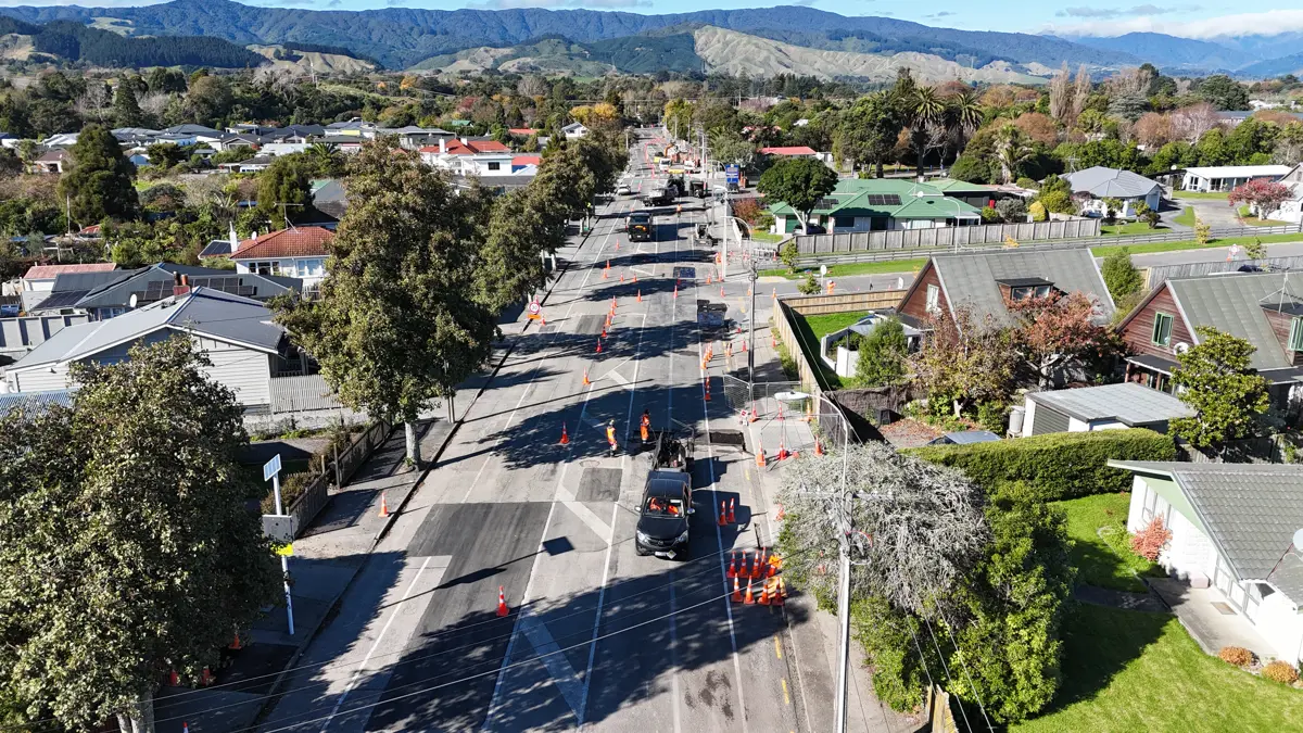 Aerial view of a suburban street undergoing roadworks, with traffic cones, construction vehicles, and workers positioned along a partially resurfaced road. Houses, mature trees, and landscaped properties line both sides of the street, while rolling hills and forested ranges form the backdrop under a clear blue sky.