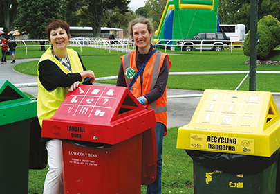 Image showing Council staff beside the waste minimisation/recycling bin hoods that can be booked through Council for use at events; in the background is a park, and people congregating