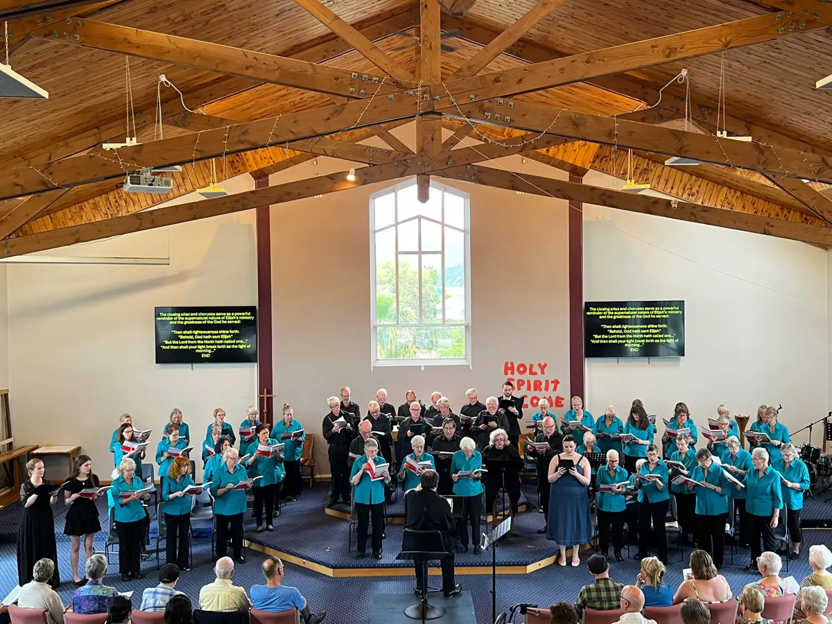 A large choir performs in front of an audience inside a church with exposed wooden rafters and a tall central window. Singers stand in rows on a raised stage around a conductor, with two display screens mounted on either side showing song lyrics. Audience members are seated in the foreground watching the performance.
