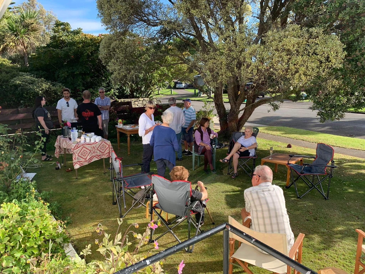 2023 Over the Fence Cuppa event in the Kāpiti community - neighbours sitting in the shade of the tree chatting, while other stand around a food table.