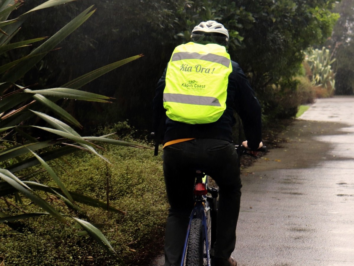A person cycling on a path with a Kāpiti Coast District Council reflective backpack cover.