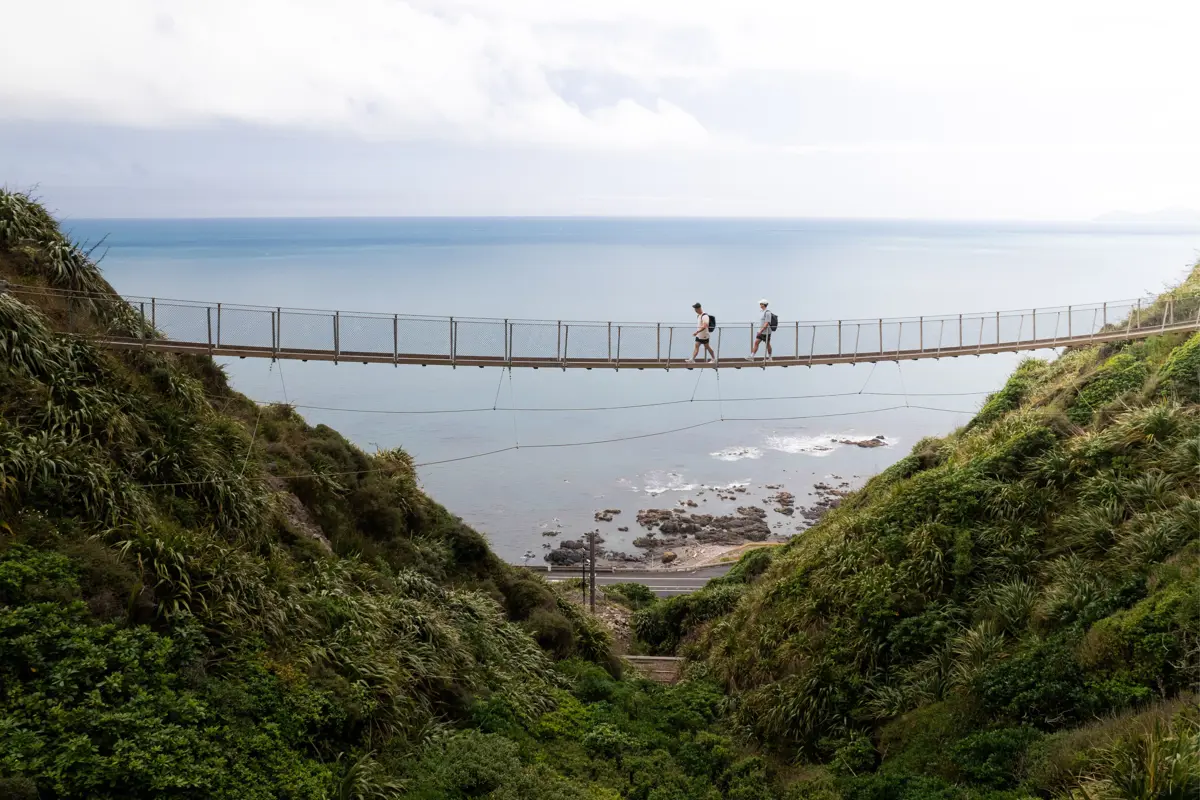Hills with the ocean in the background and a bridge stretching between hills with two hikers crossing the bridge