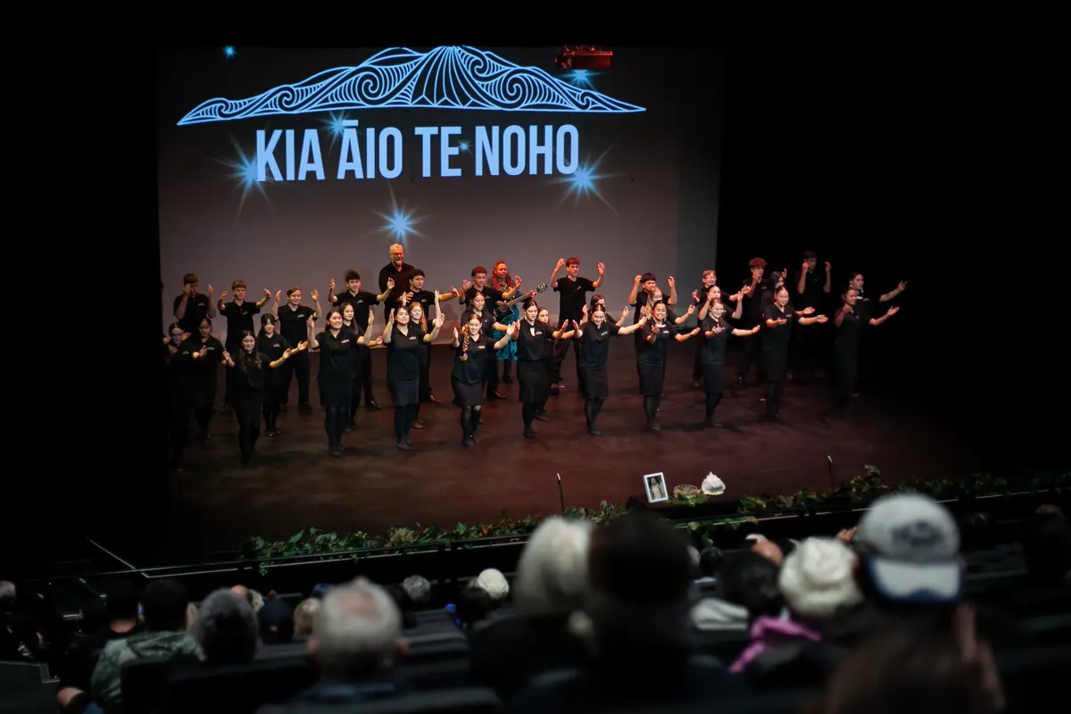 Performers dressed in black stand on a theatre stage presenting a group performance in front of a large illuminated backdrop displaying the words “KIA ĀIO TE NOHO” beneath a stylised mountain design. Audience members are visible in the foreground, watching from theatre seating.