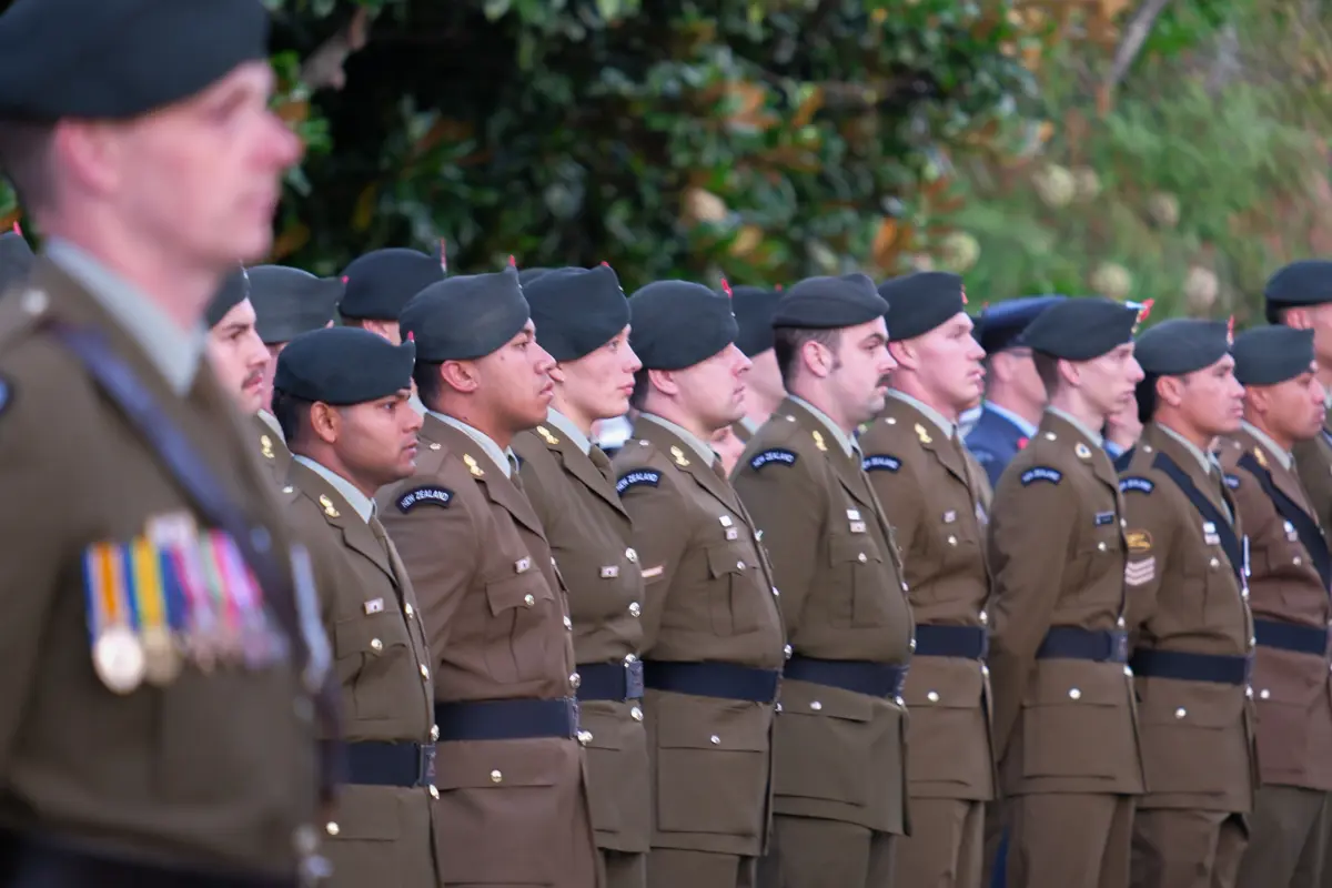A row of uniformed New Zealand Army personnel wearing berets stand in formation outdoors, viewed side‑on, with medals and insignia visible on their uniforms.