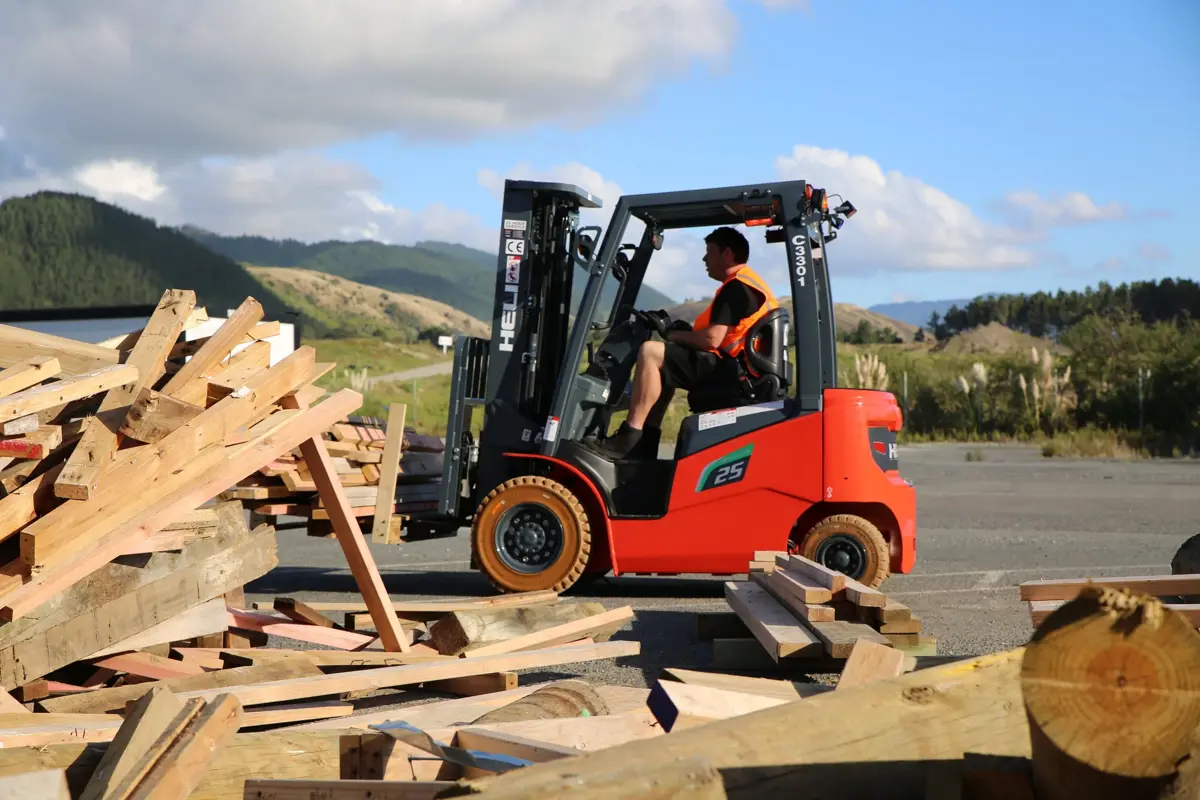 A red forklift operated by a worker wearing a high-visibility vest moves through a timber recycling area, surrounded by stacks and piles of scrap wood. The scene is set outdoors with rolling green hills, trees, and a partly cloudy blue sky in the background. Sunlight highlights the timber and machinery, showing materials being sorted or transported at a waste recovery site.