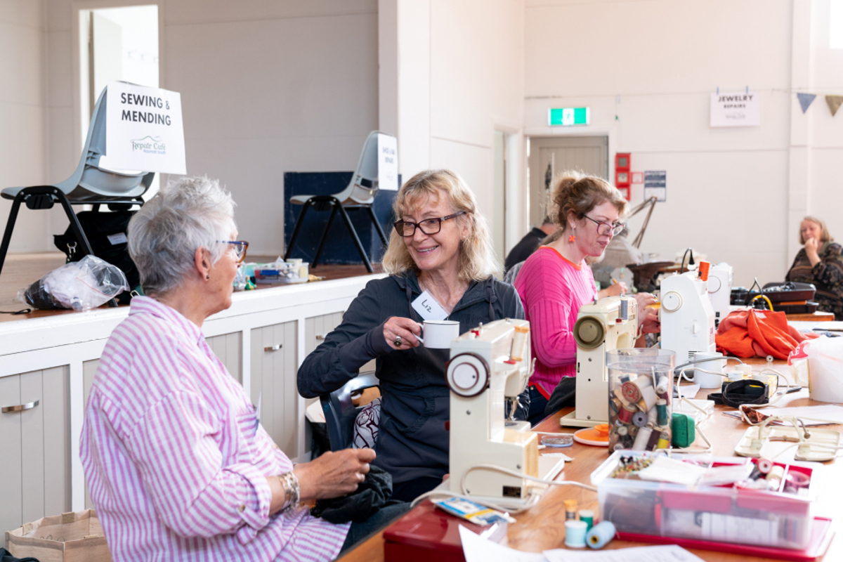 Two ladies sitting together chatting at the Raumati south repair cafe. One is holding a cup of tea. There are sewing machines on the tables that they are sitting at