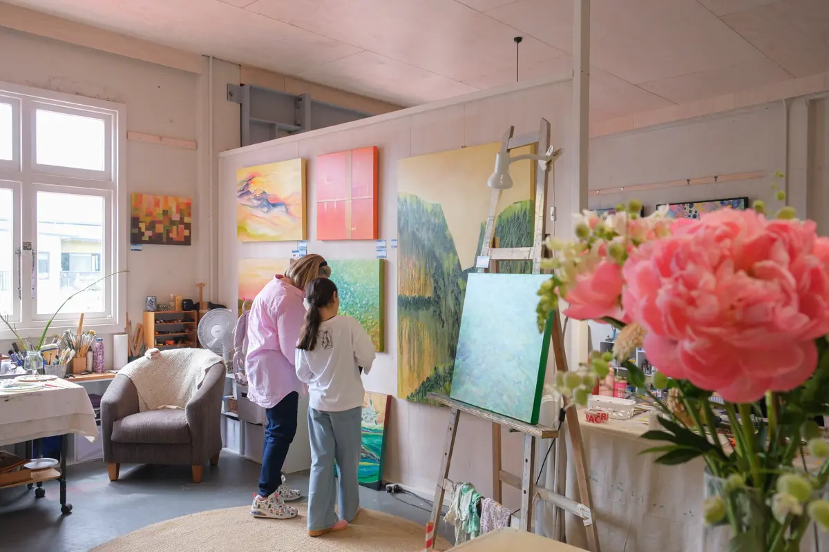 Visitor viewing colourful landscape and abstract paintings displayed inside a bright artist’s studio. An easel with a large canvas stands among artwork, painting supplies and a vase of pink flowers in the foreground.