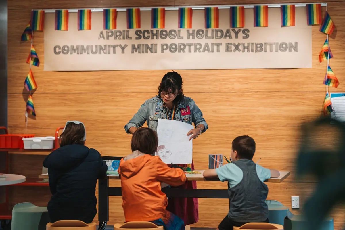 Librarian running a art workshop with three children in front of her. In the background there are rainbow flags surrounding a big sign that reads "April school holidays Community mini portrait exhibition"