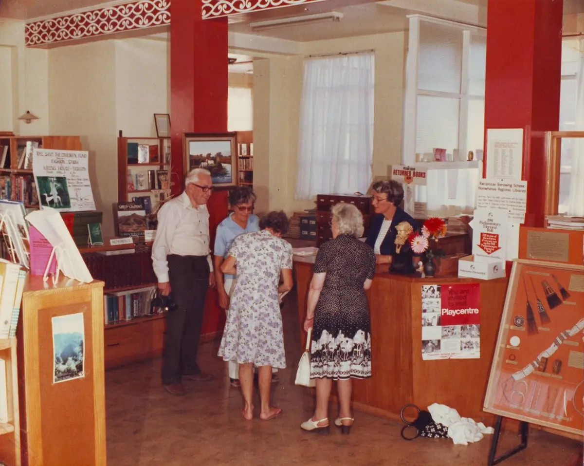 Vintage photo of Ōtaki library interior showing borrowers at issues desk gathered talking.