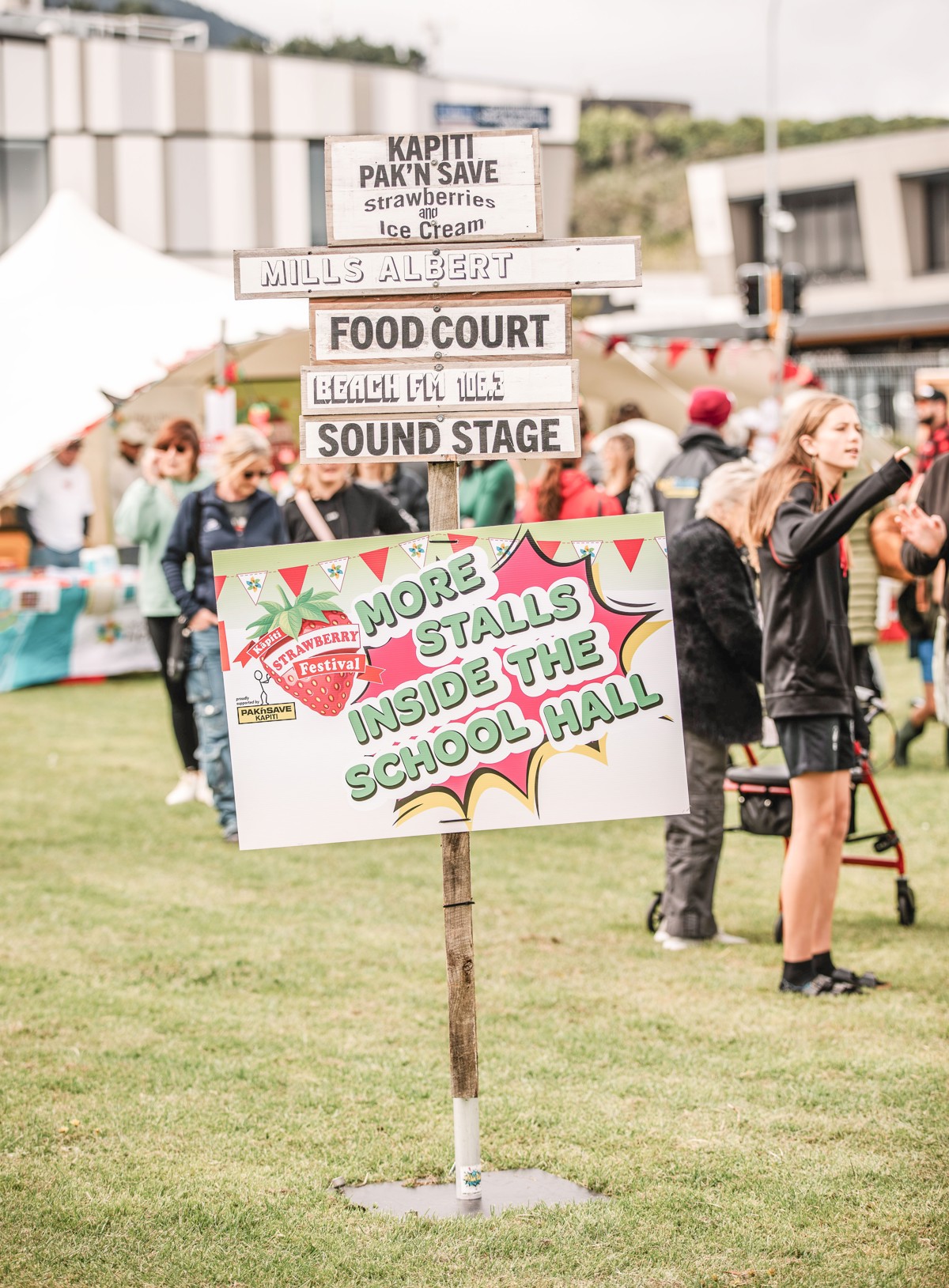 Signs at Kāpiti Strawberry Festival including Pak n Save strawberries and cream, Mills Albert Food Court, Beach FM Sound Stage and more stalls inside the school hall.