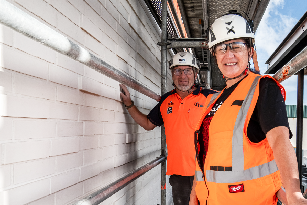 Alt text: Two people wearing hard hats, safety glasses, and high-visibility vests stand beside a newly completed light-coloured brick wall at a construction site. Scaffolding surrounds the area, with blue sky visible in the background.