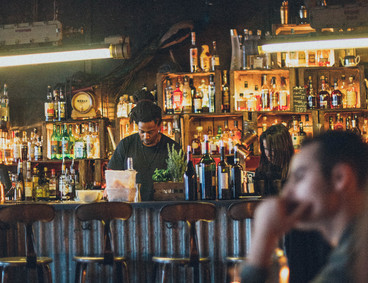 Photo showing a bar, with alcohol on shelves, two bartenders working, and a man in the foreground, slightly out of focus