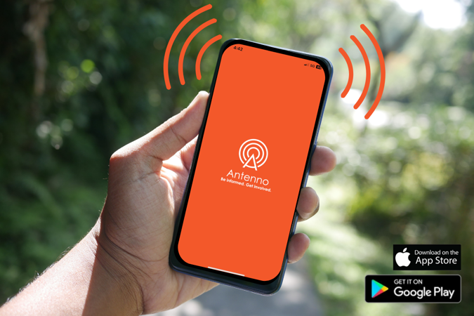 Photo of a hand holding a smartphone with the Antenno opening screen displayed. The bottom right corner of the photo has the "Download on the App Store" and "Get it on Google Play" logos.