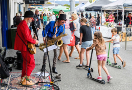 Photo of two musicians busking at the Paraparaumu Saturday Market, with children scooting past on the footpath.