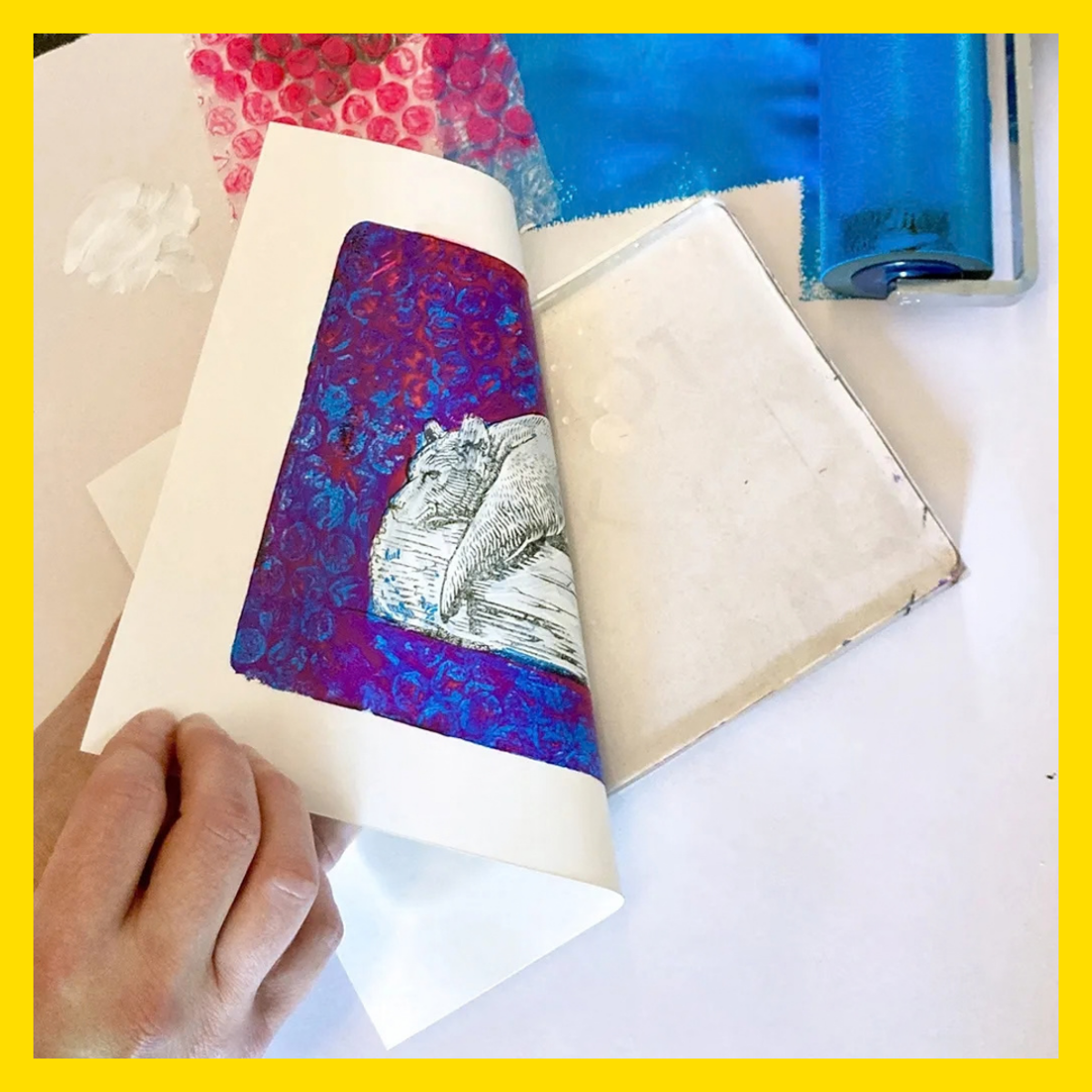 A hand lifts a sheet of paper to reveal a colourful gel print in progress. The print features a vivid purple and blue patterned background with a black-and-white illustration of a resting bear. Art materials are scattered around the workspace, including bubble wrap, acrylic paint, and a blue brayer roller used for printmaking. The image highlights a creative mixed-media printing process.