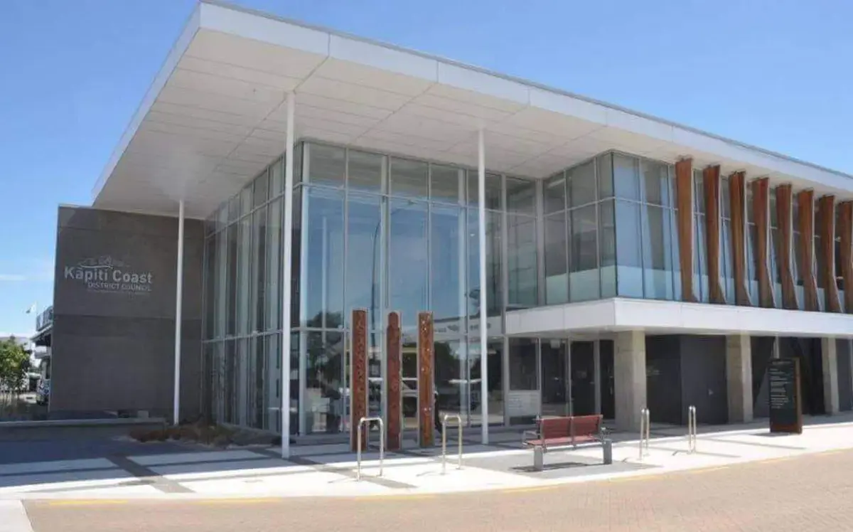 Modern Kāpiti Coast District Council building with large glass walls and a wide white roof overhang. The entrance features tall wooden panels and glass doors, with bike stands and a bench out front. The building sits beside a paved street under a clear blue sky.