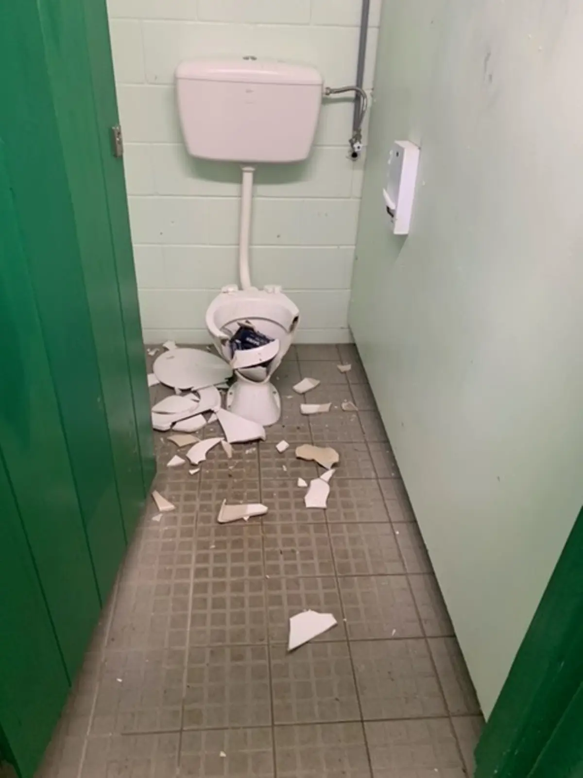 Public toilet cubicle with a broken toilet bowl and seat, shattered ceramic pieces scattered across the tiled floor. Pale green walls and a cistern mounted above the damaged toilet are visible.