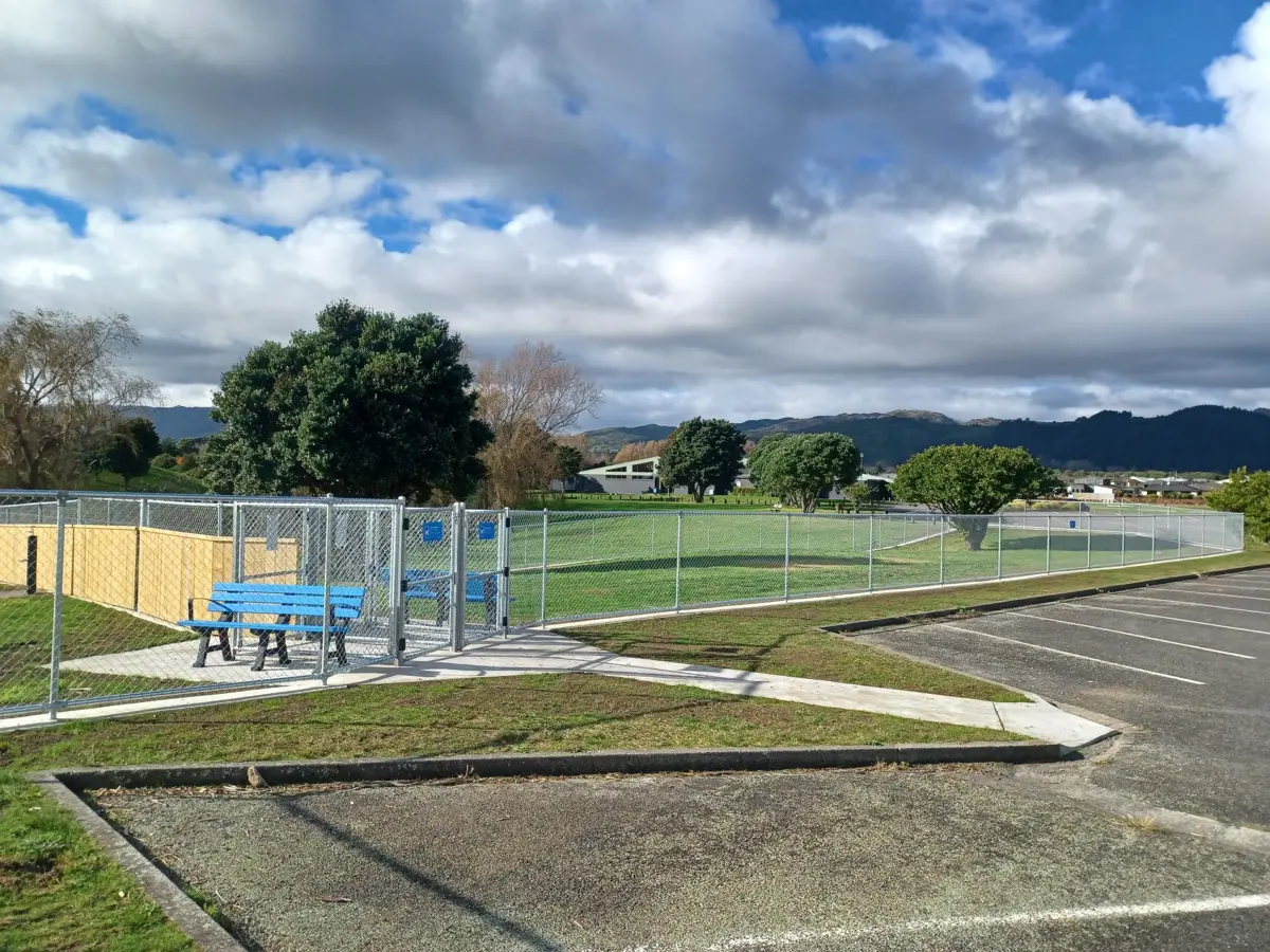 A fenced outdoor dog exercise or play area sits beside an empty car park, with a bright blue bench near the gate and grassy fields and trees under a partly cloudy sky, with low hills in the background.