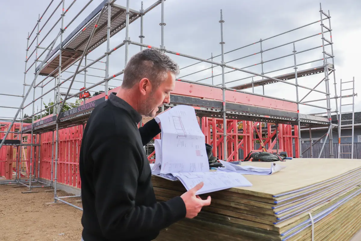Person reviews building plans on stacked boards at an active construction site, with red timber framing and metal scaffolding surrounding a partially built structure under overcast skies.