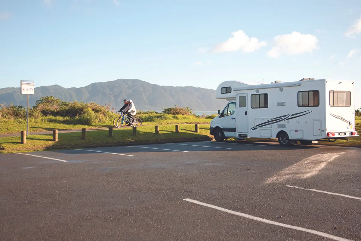 Campervan parked in a freedom camping site along Marine Parade, Paraparaumu. A couple of cyclists ride along the path and Kāpiti Island is in the background.