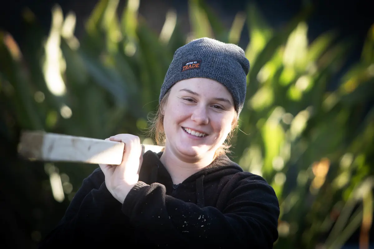 Person holds a timber beam over shoulder, wearing a beanie, outdoors with greenery in the background and soft sunlight highlighting the scene.