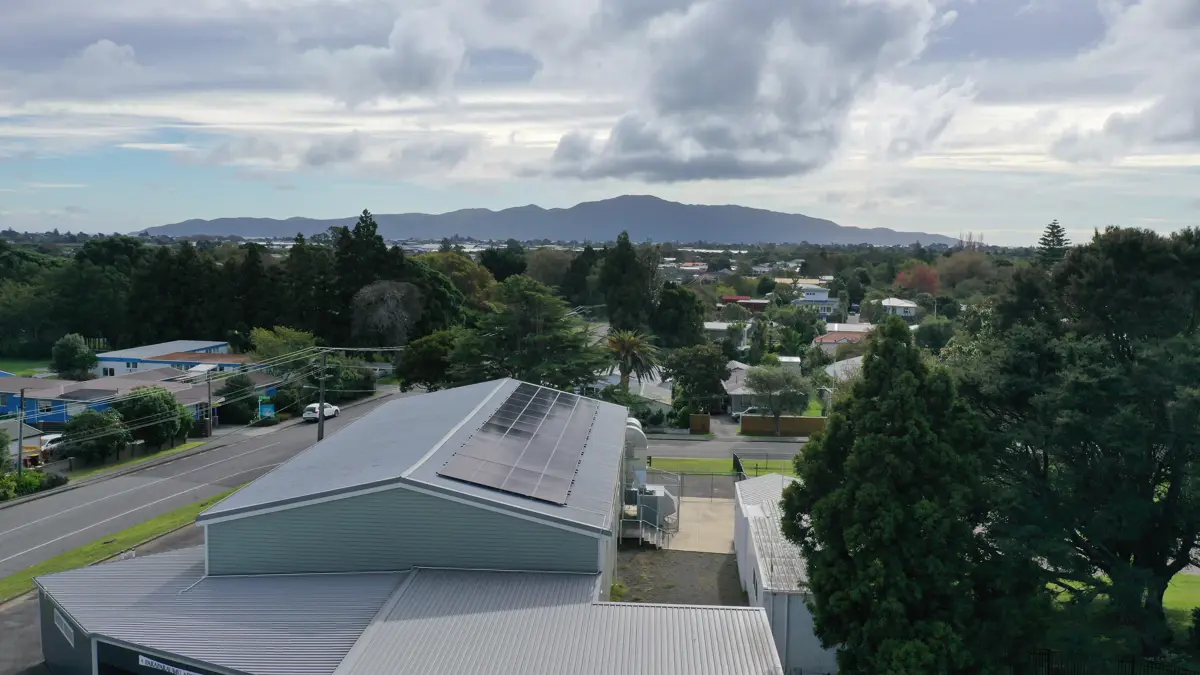 Aerial view of Paraparaumu Memorial Hall with solar panels on the roof, surrounded by trees and nearby houses, with Kāpiti Island in the distance under a cloudy sky