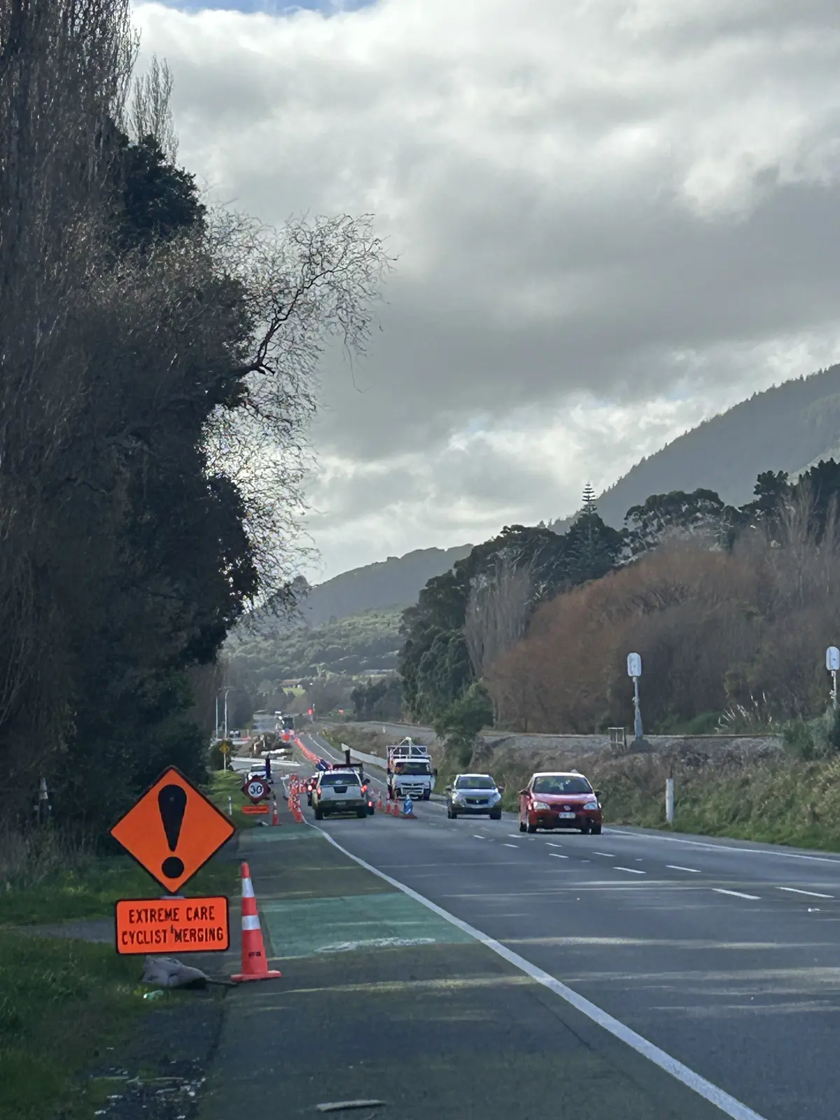 Roadworks underway on a roadside corridor with traffic cones, temporary speed restrictions and traffic management vehicles. A warning sign reading “Extreme Care Cyclist Merging” is positioned beside the road, with cars travelling through the work area and hills in the background.