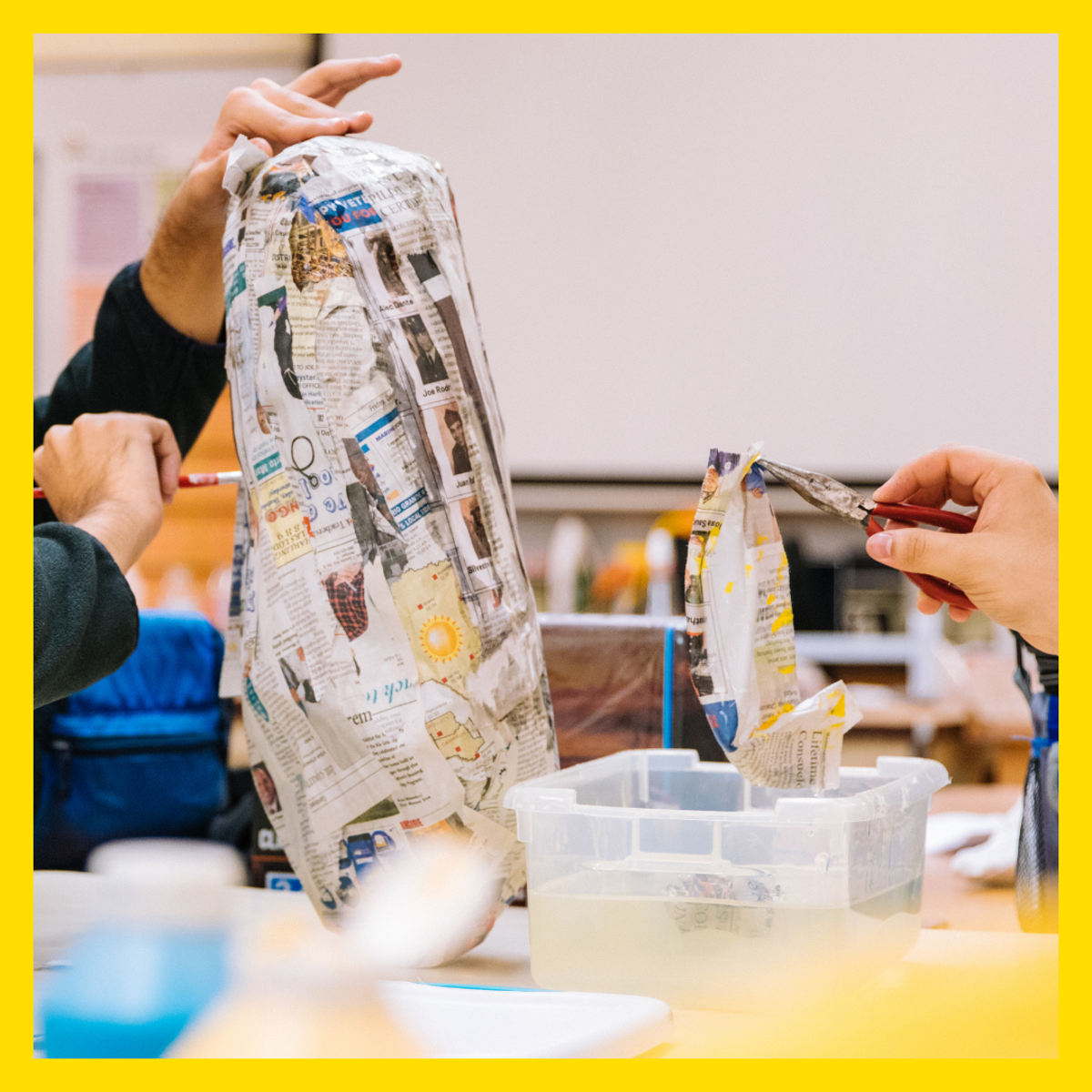 Hands working with paper‑mâché, applying strips of newspaper to a tall paper form and dipping pieces into a container of paste on a craft table.