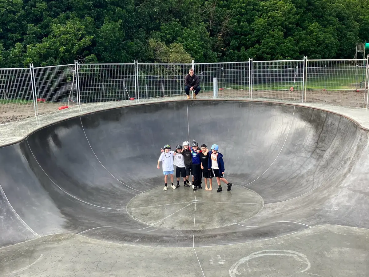 A large concrete skate bowl at a skatepark, viewed from above. Six young skaters wearing helmets stand together in the centre of the bowl, while another person sits on the edge overlooking the scene. Temporary fencing surrounds the skate area, with dense green trees in the background. The skate bowl appears newly constructed and is free of skateboards or scooters.