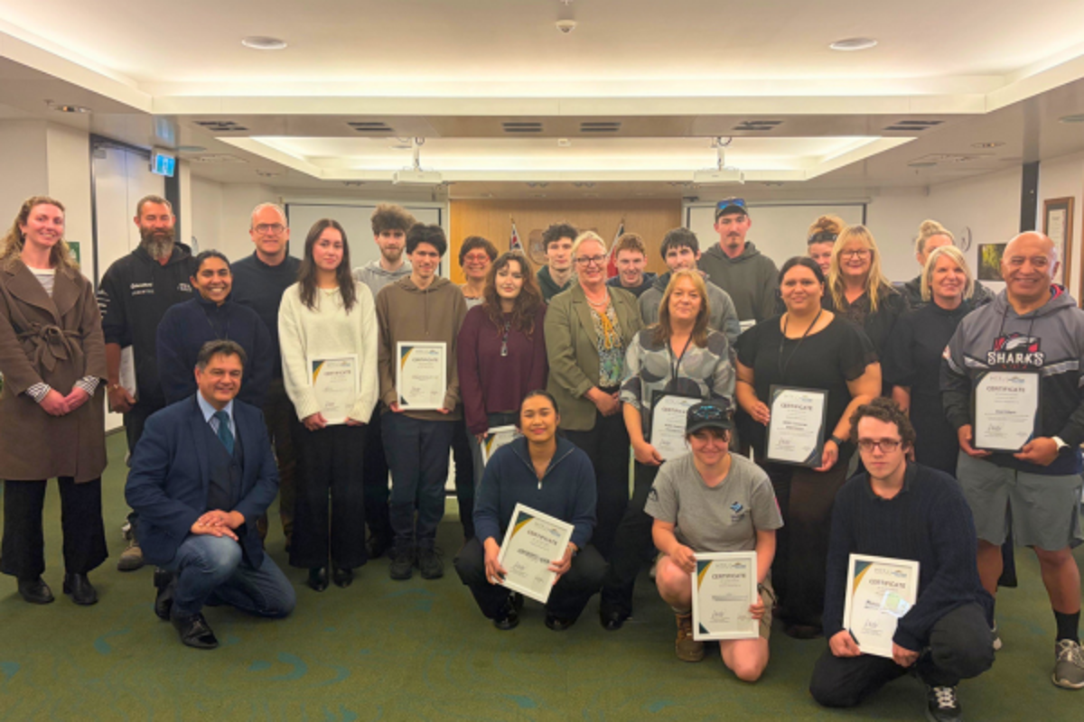 Group photo inside a council chamber or meeting room, with more than 20 people gathered for a certificate presentation. Several people hold certificates while standing and kneeling in rows. Tables, presentation screens, and council insignia are visible in the background.