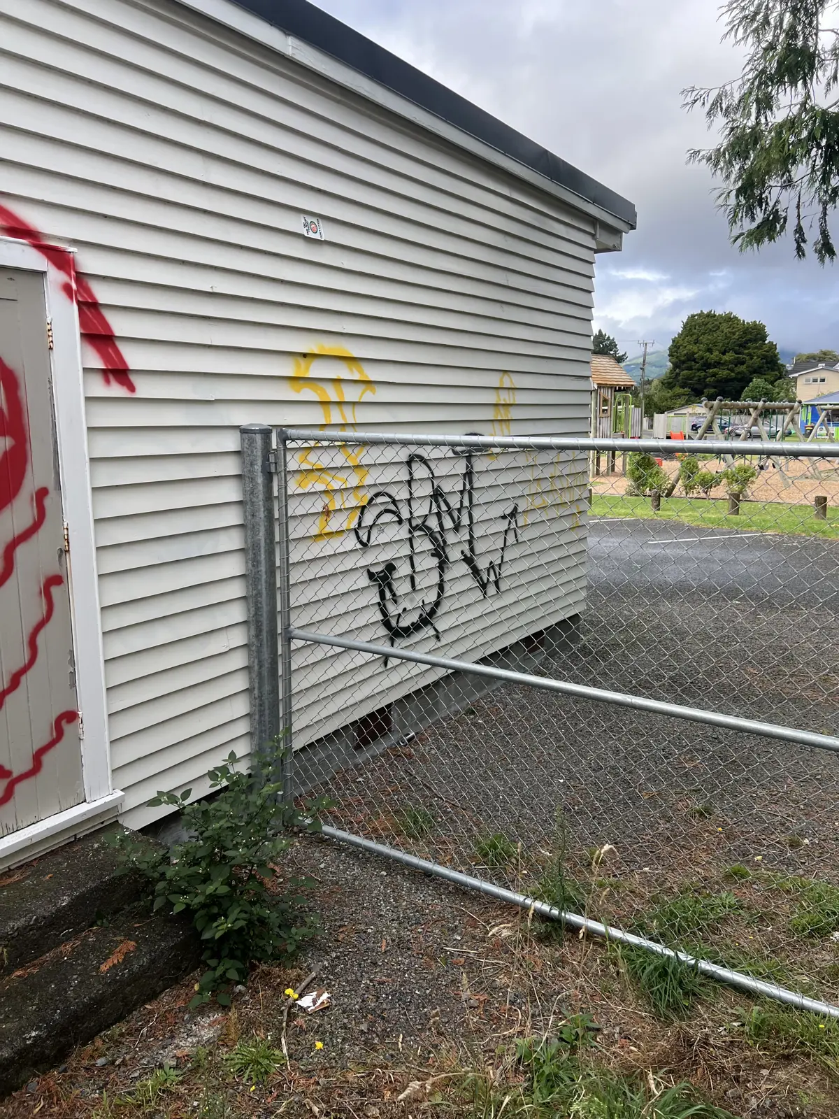 Exterior wall of a light-coloured community hall marked with spray-painted graffiti in red, yellow and black. A chain-link fence runs alongside the building, with a playground and trees visible in the background under a cloudy sky.